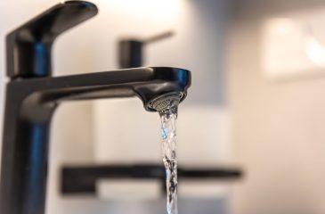 Macro of a black faucet aerator with a ribbon of water. Focus on metal texture and cleanliness for a modern bathroom.