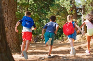 Rear View Of Group Of Children Running Along Trail Through Forest On Hiking Adventure In Countryside