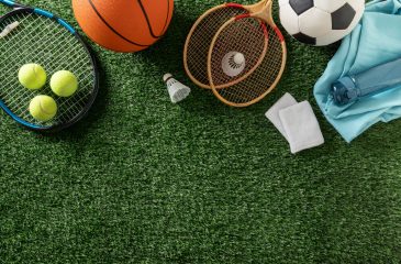 Sports equipment with tennis, badminton, basketball and soccer gear arranged on green grass, top view with large copy space.